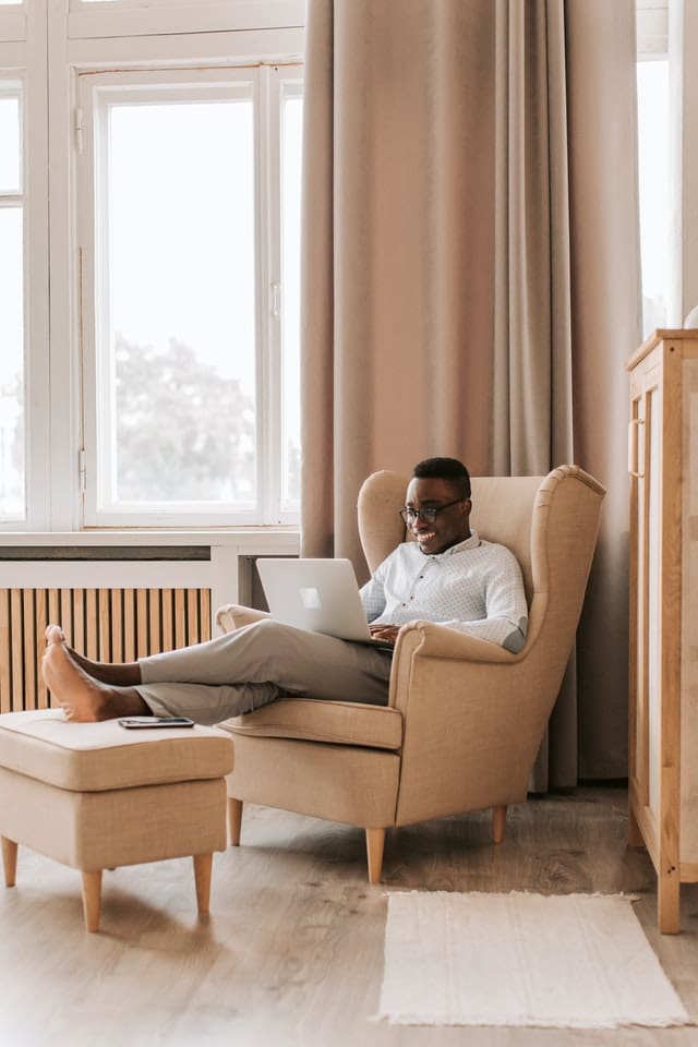 Young man sitting next to a window smiling at something on his laptop